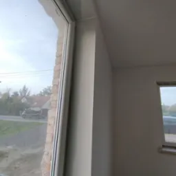 Świeżo wyszpachlowana ściana wewnętrzna w trakcie prac wykończeniowych, widoczne niedoskonałości i nierówności przed szlifowaniem, okno z widokiem na ulicę i zabudowania w tle.