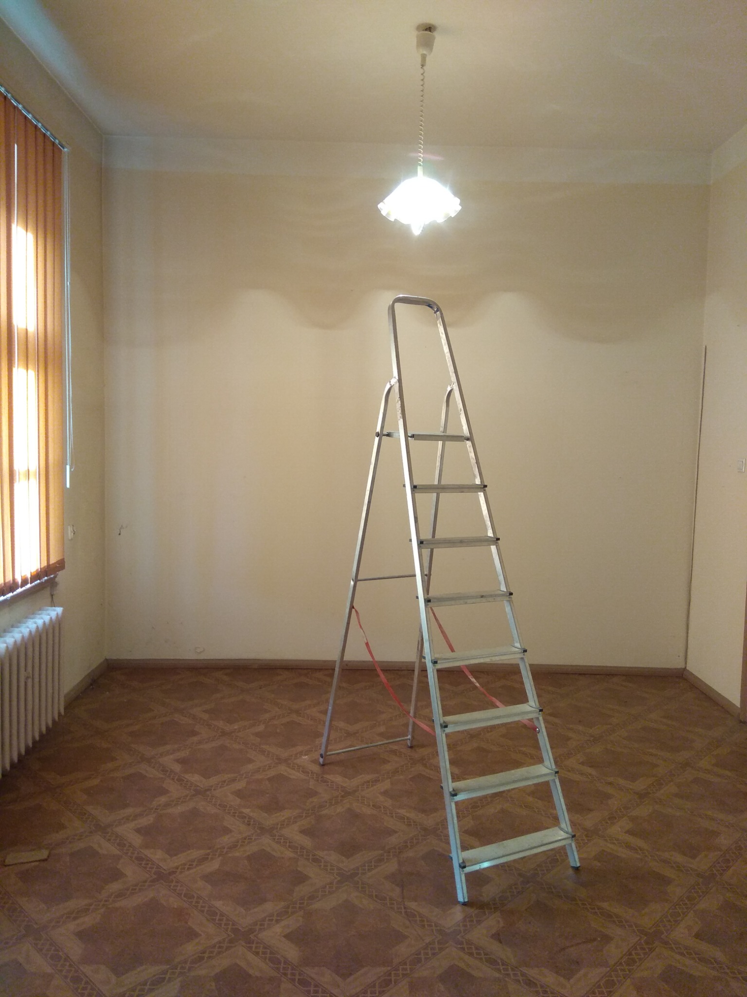 Pokój przed remontem z aluminiową drabiną rozstawną na środku, beżowe ściany przygotowane do malowania, wzorzysta podłoga, grzejnik pod oknem z żaluzjami.