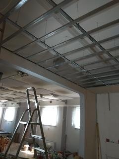 Stelaż sufitu podwieszanego z płyt kartonowo-gipsowych, widoczna drabina aluminiowa, wnętrze w trakcie prac wykończeniowych, z widocznymi instalacjami i oknami w tle.