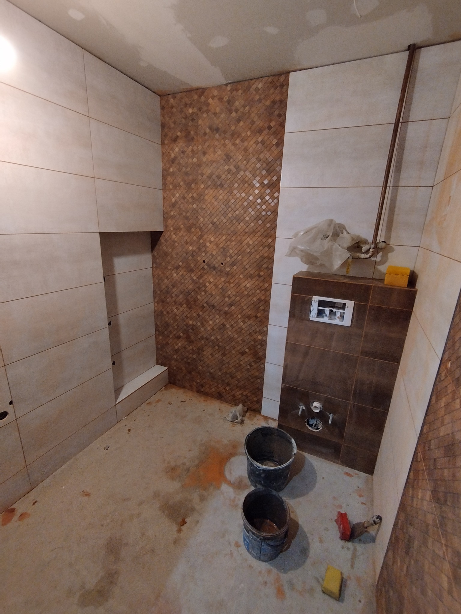 Wnętrze łazienki w trakcie kładzenia płytek, widoczne dwa rodzaje płytek: beżowe duże panele oraz mozaika w odcieniach brązu, na podłodze stoją dwa wiadra i gąbka, ściany częściowo wykończone...