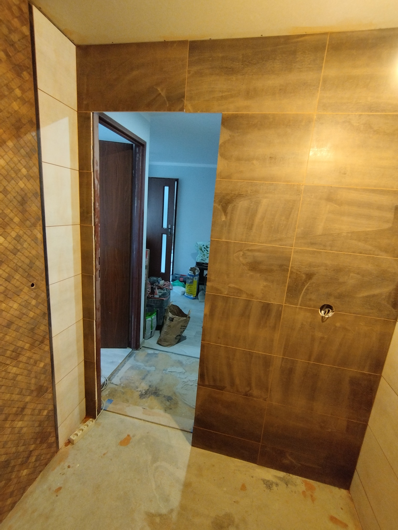 Układanie brązowych płytek imitujących drewno w łazience, widoczne narzędzia i akcesoria budowlane w tle, surowy stan podłogi przed fugowaniem.