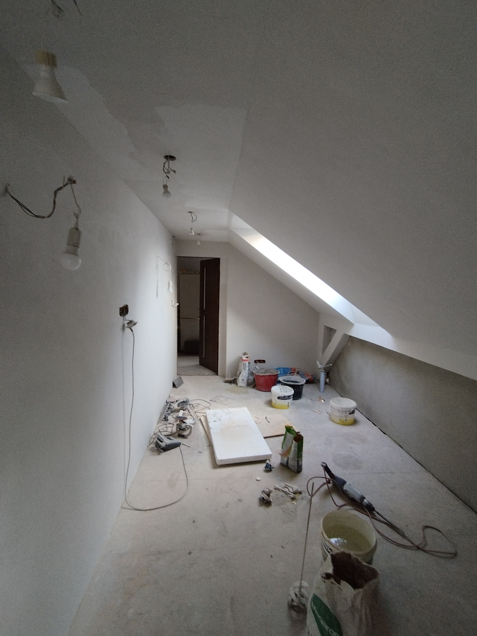Poddasze w trakcie szpachlowania ścian i sufitu, widoczne narzędzia, wiadra z zaprawą, kable elektryczne i niedokończone oświetlenie.
