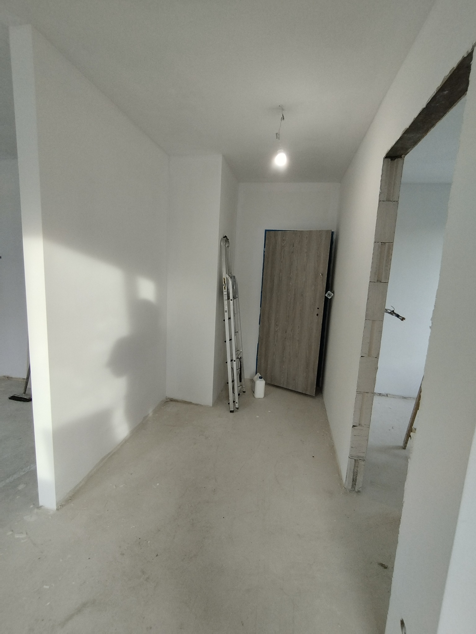 Wnętrze korytarza w trakcie wykańczania: świeżo pomalowane białe ściany, surowy betonowy podkład, częściowo zamontowane drzwi w odcieniach szarości, drabina oparta o ścianę, widoczna żarówka...