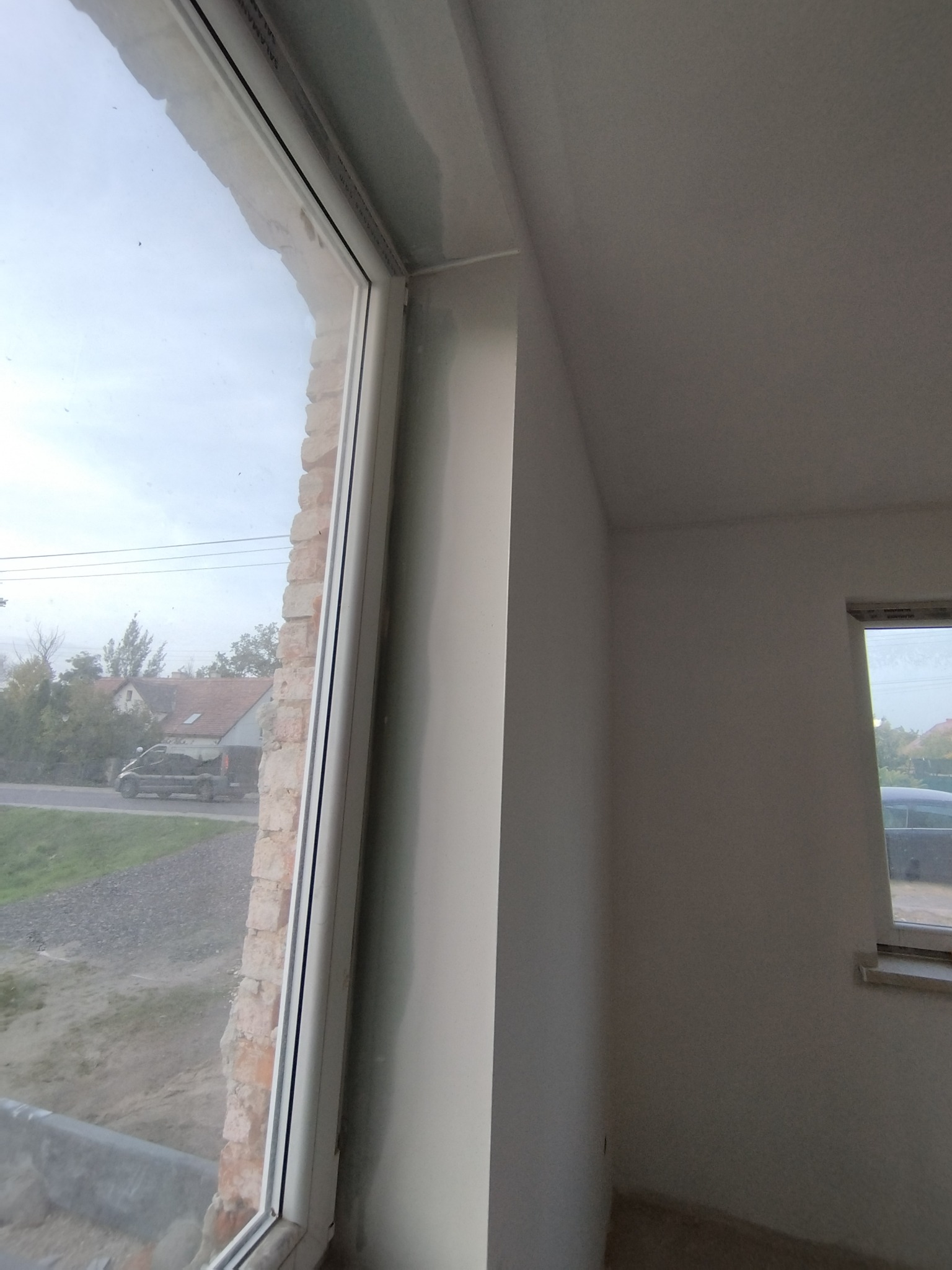 Świeżo wyszpachlowana ściana wewnętrzna w trakcie prac wykończeniowych, widoczne niedoskonałości i nierówności przed szlifowaniem, okno z widokiem na ulicę i zabudowania w tle.