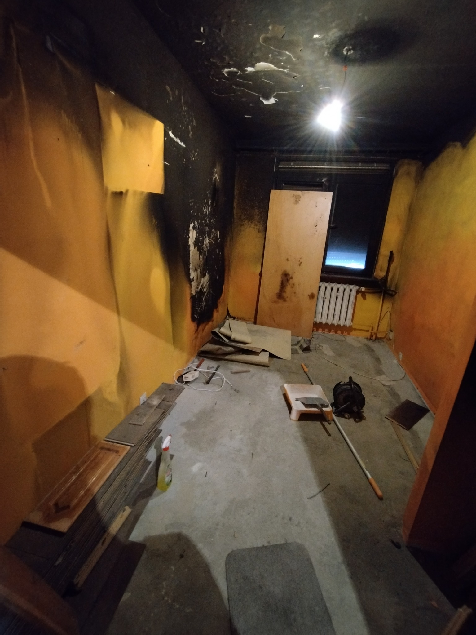 Pokój w trakcie remontu: częściowo pomalowane na żółto ściany, ślady po pożarze na suficie i ścianie, panele podłogowe, drabinka malarska i narzędzia malarskie na podłodze.