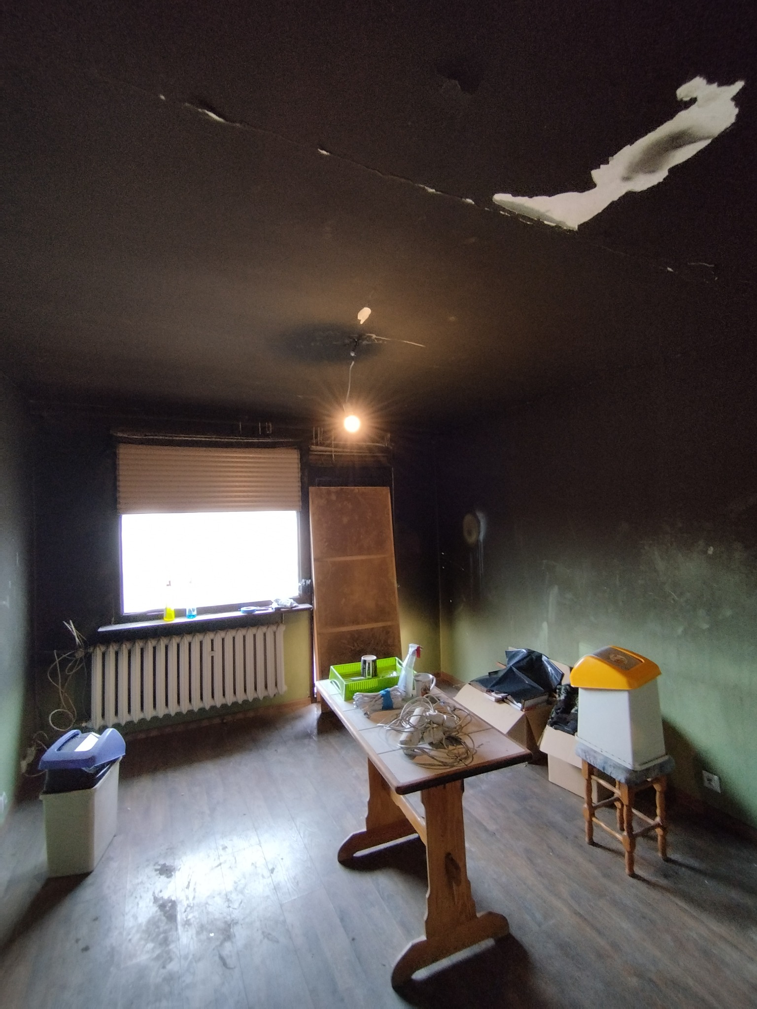 Pokój w trakcie przygotowań do remontu: ciemne ściany z widocznymi śladami, odchodząca farba na suficie, drewniany stół z narzędziami i akcesoriami, okno z żaluzjami i grzejnikiem.