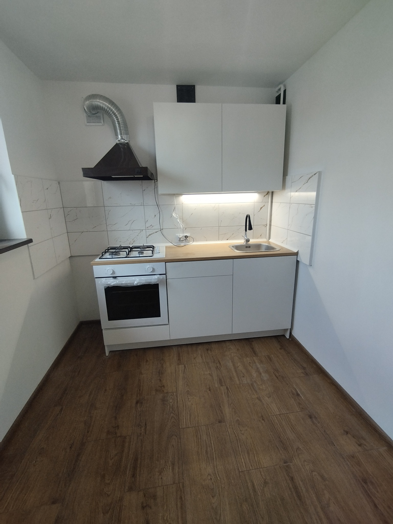 Nowoczesna, minimalistyczna kuchnia w odcieniach bieli i brązu z drewnianą podłogą, białymi szafkami, płytą gazową z piekarnikiem, czarnym okapem i marmurkowymi płytkami na ścianie.