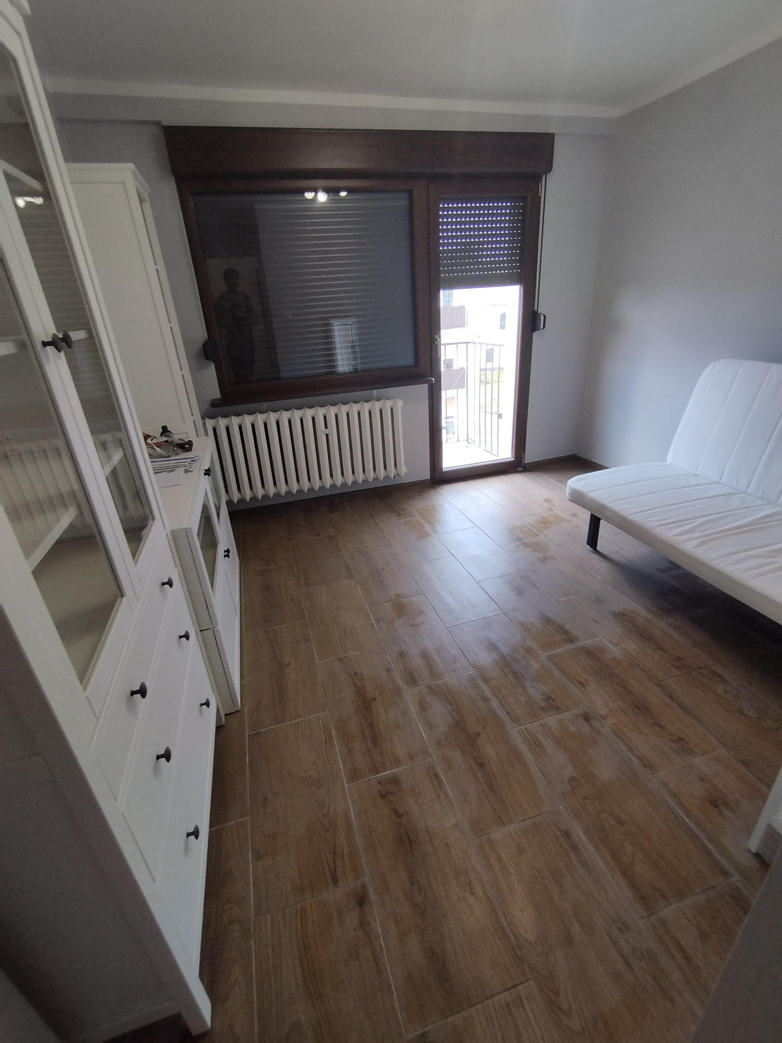 Ułożone panele podłogowe imitujące drewno w pokoju z białymi meblami i grzejnikiem pod oknem.