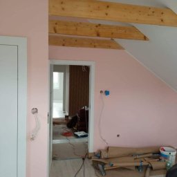 GREEN BUD ARTUR ZIELIŃSKI - Różowa ściana w trakcie malowania, widoczne drewniane belki stropowe, zabezpieczona podłoga i wystające kable elektryczne