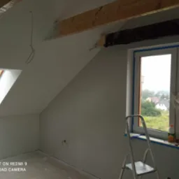 Poddasze po świeżym malowaniu na szaro, z oknem dachowym i zabezpieczonym oknem pionowym, widoczne drewniane belki stropowe i drabina malarska.