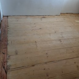 GREEN BUD ARTUR ZIELIŃSKI - Fragment pokoju z drewnianą podłogą z desek i fragmentem ściany z widocznymi śladami po szpachlowaniu.