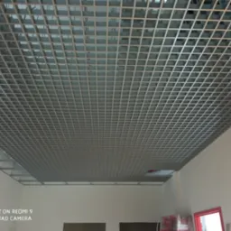 Metalowa sufit podwieszany w kratkę, widoczne elementy konstrukcyjne i białe ściany pomieszczenia.