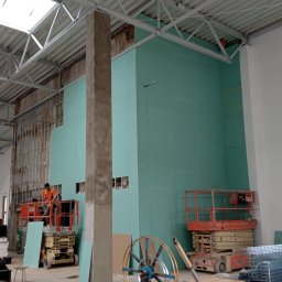 GREEN BUD ARTUR ZIELIŃSKI - Montaż płyt karton gips na ścianie w hali produkcyjnej, widoczny pracownik na podnośniku, metalowa konstrukcja sufitu i narzędzia budowlane na podłodze.