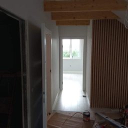 GREEN BUD ARTUR ZIELIŃSKI - Korytarz w trakcie odświeżania: pomalowane ściany, drewniane belki pod sufitem, pionowe panele dekoracyjne na ścianie, zabezpieczona podłoga, widoczne narzędzia i puszka z farbą.