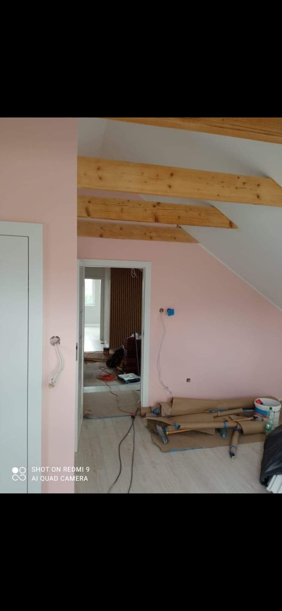 Różowa ściana w trakcie malowania, widoczne drewniane belki stropowe, zabezpieczona podłoga i wystające kable elektryczne