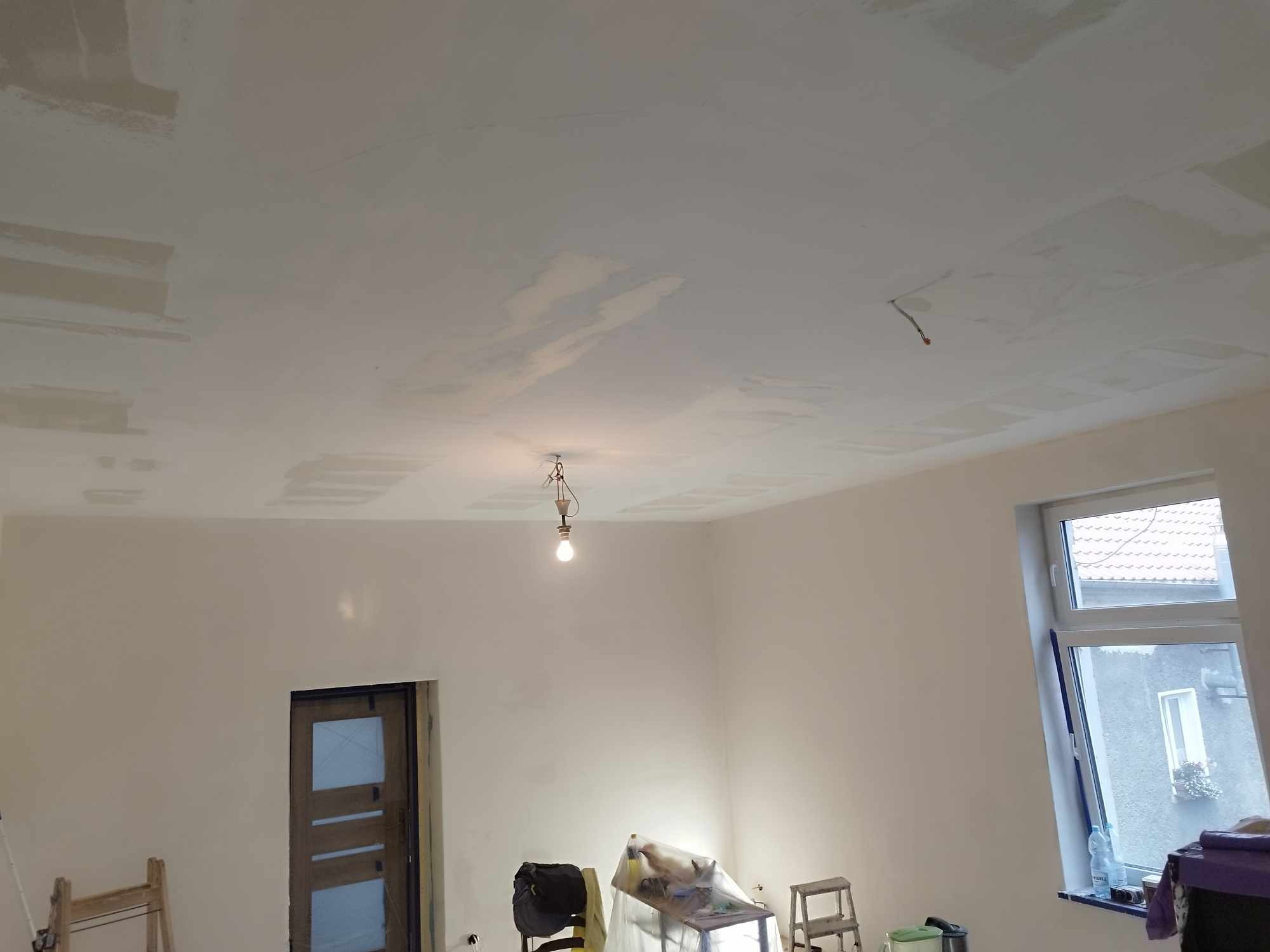 Pokój w trakcie remontu: sufit z widocznymi łatami gładzi, ściany przygotowane do malowania, widoczne narzędzia i drabiny, surowa żarówka na kablu.