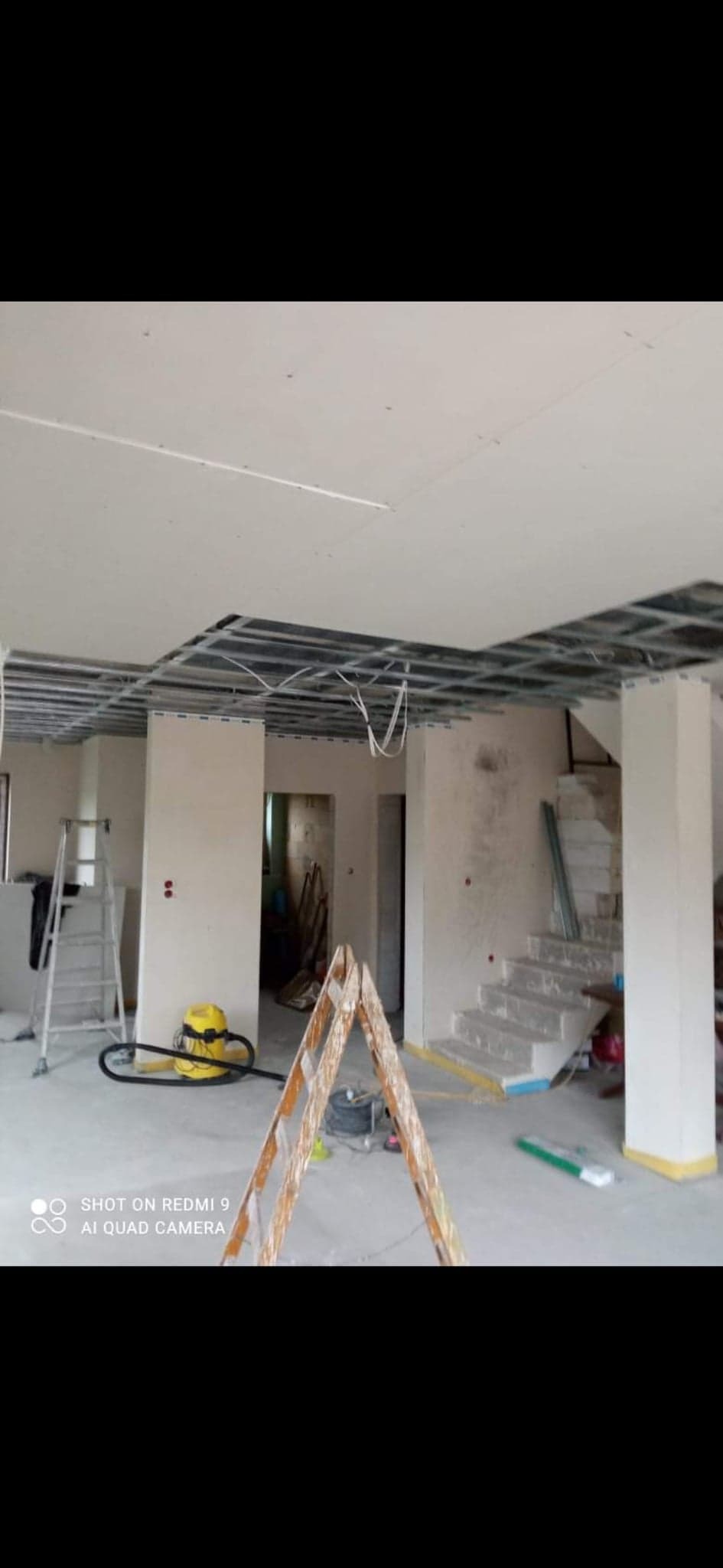 Wnętrze w trakcie montażu płyt kartonowo-gipsowych: widoczne metalowe profile sufitowe, częściowo zamontowane płyty, drabina, odkurzacz przemysłowy i schody w surowym stanie.