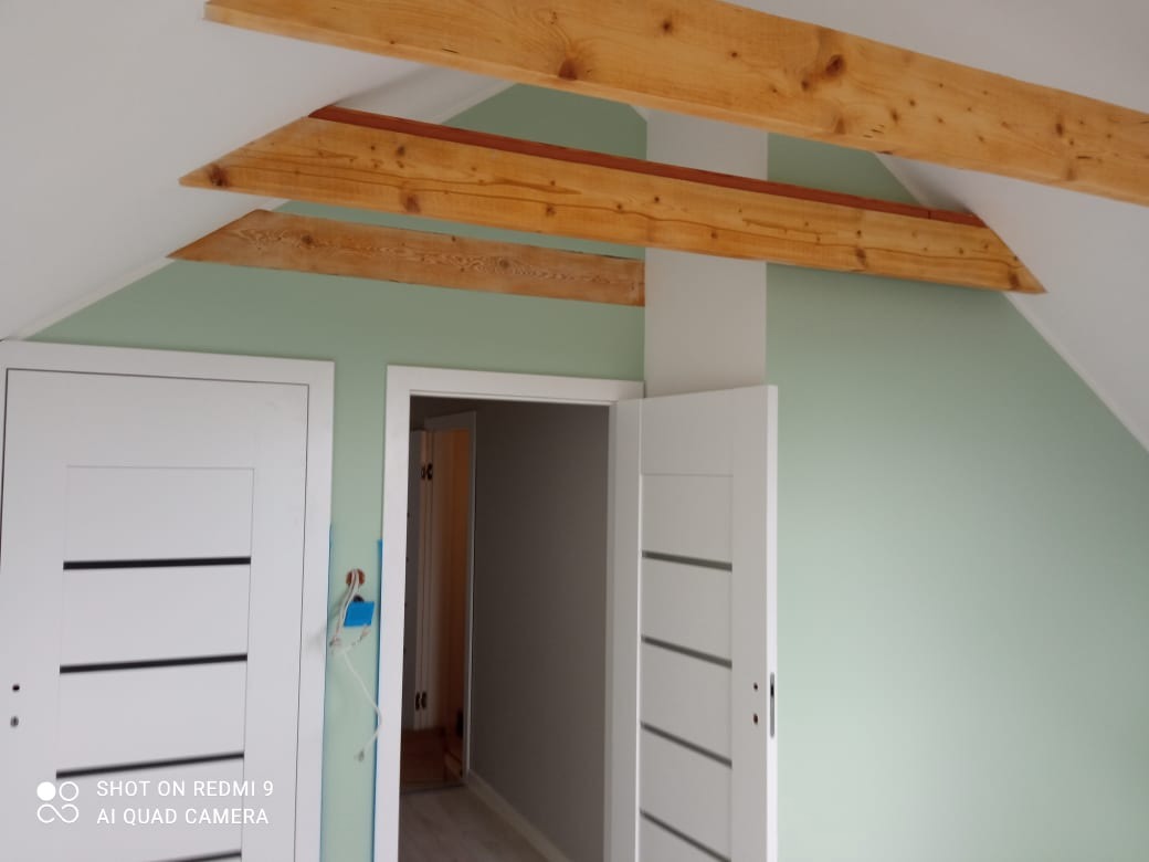 Poddasze po remoncie: świeżo pomalowane ściany w pastelowym odcieniu zieleni, drewniane belki stropowe, białe drzwi z poziomymi frezami, widoczne ślady po instalacji elektrycznej.