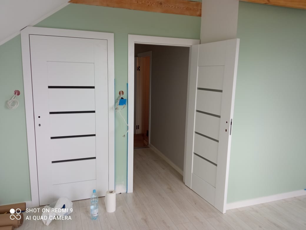 Dwie białe, nowoczesne drzwi wewnętrzne z poziomymi, ciemnymi paskami, jedna zamknięta, druga uchylona, na tle świeżo pomalowanych, jasnych ścian w odcieniu zieleni, widoczne ślady po pracach...