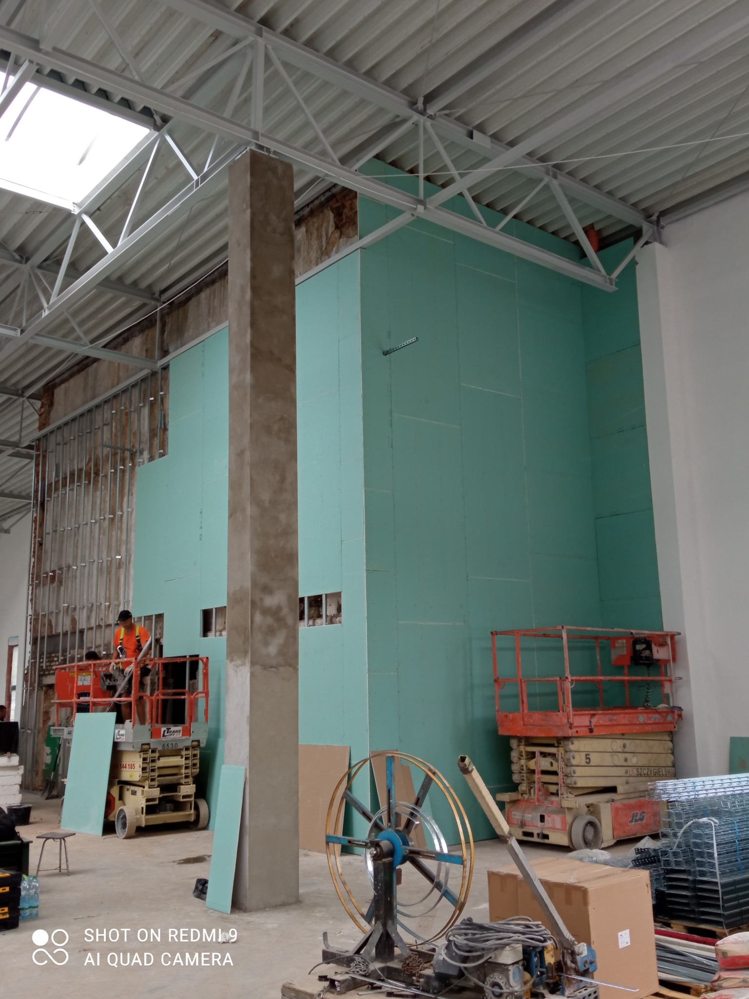 Montaż płyt karton gips na ścianie w hali produkcyjnej, widoczny pracownik na podnośniku, metalowa konstrukcja sufitu i narzędzia budowlane na podłodze.