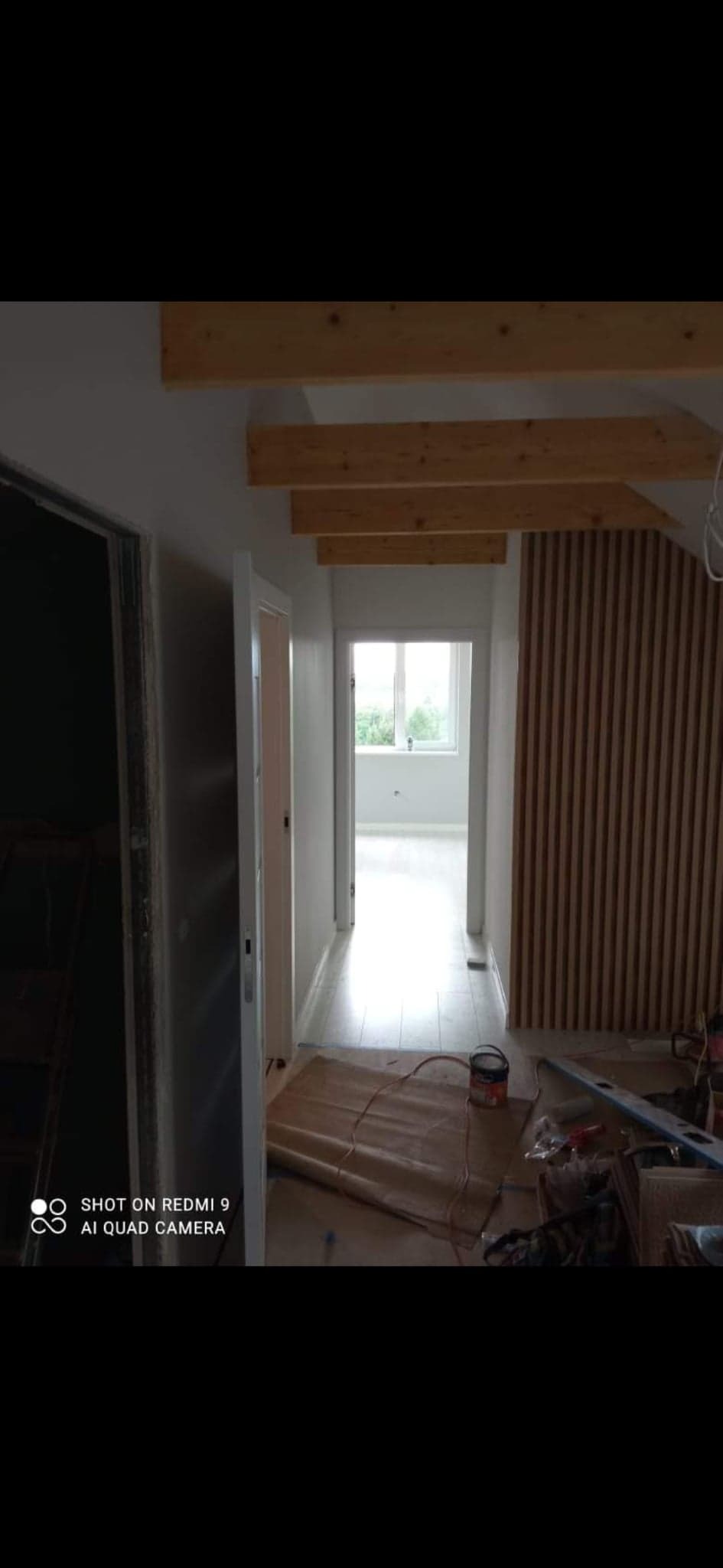 Korytarz w trakcie odświeżania: pomalowane ściany, drewniane belki pod sufitem, pionowe panele dekoracyjne na ścianie, zabezpieczona podłoga, widoczne narzędzia i puszka z farbą.