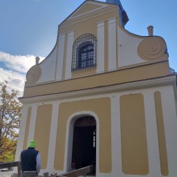 Matmar System - Odrestaurowana fasada kościoła w odcieniach kremu i ochry, widok z dołu na pracownika w zielonej czapce na tle nieba i rusztowania.