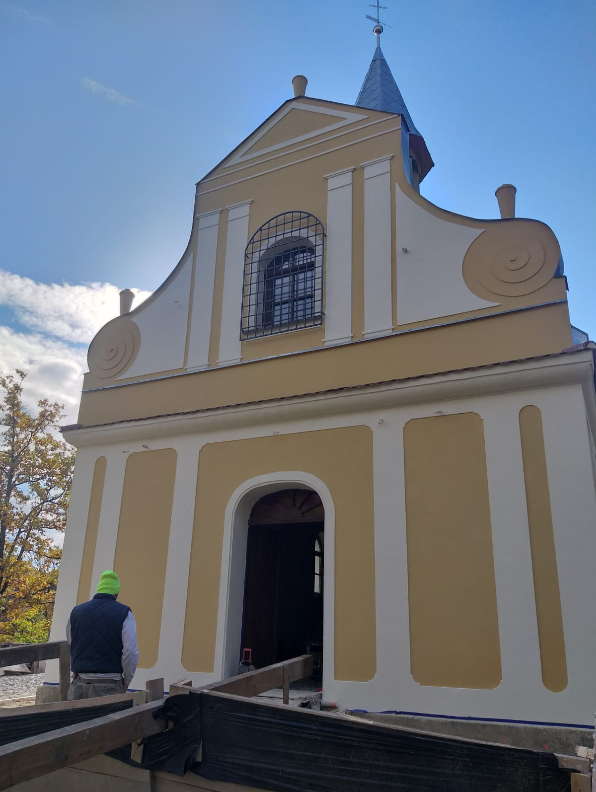 Odrestaurowana fasada kościoła w odcieniach kremu i ochry, widok z dołu na pracownika w zielonej czapce na tle nieba i rusztowania.