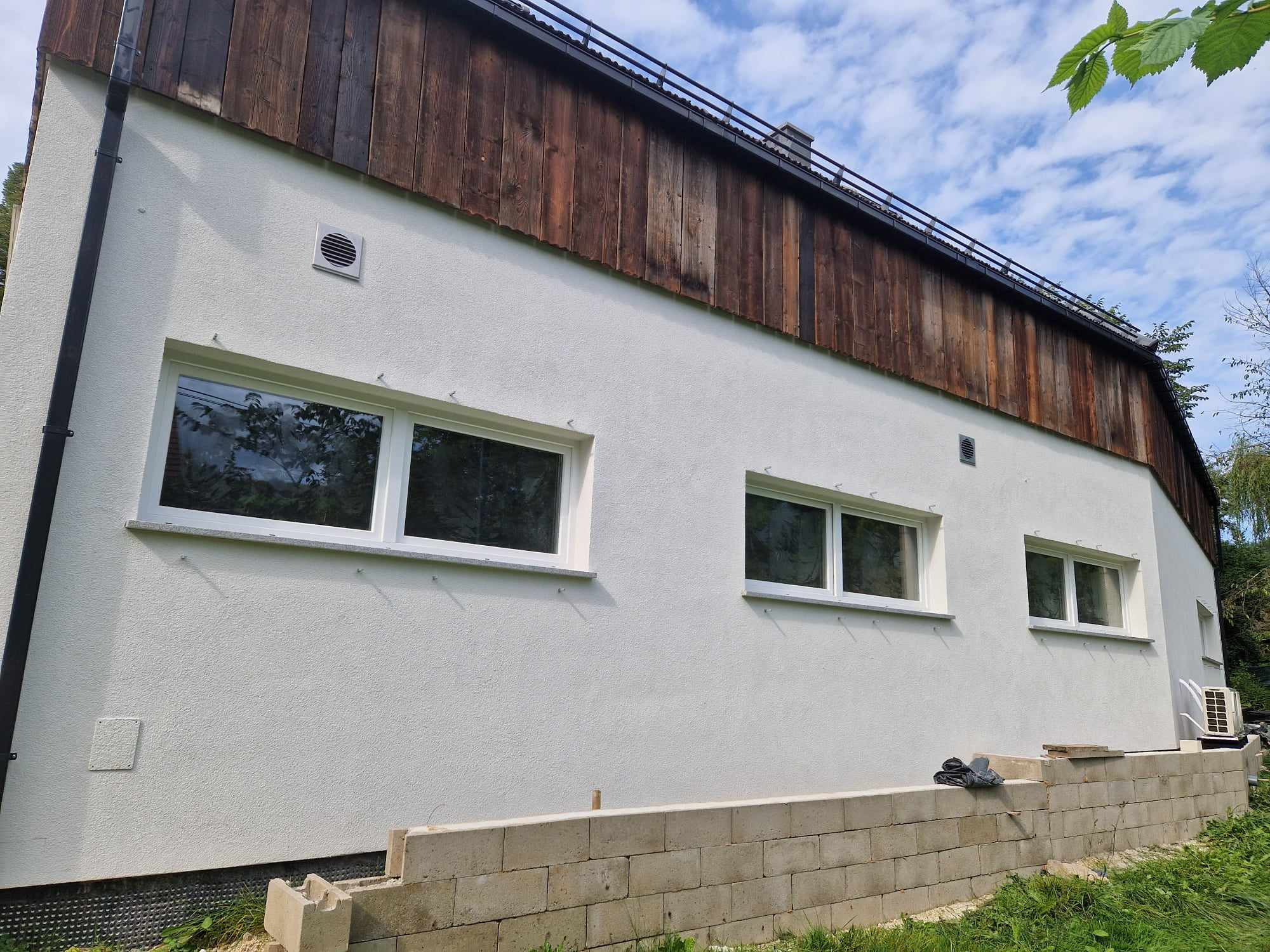 Boczny widok domu z białą elewacją o chropowatej fakturze, drewnianą górną częścią i białymi oknami, widoczny fragment fundamentu z bloczków betonowych i zieleni