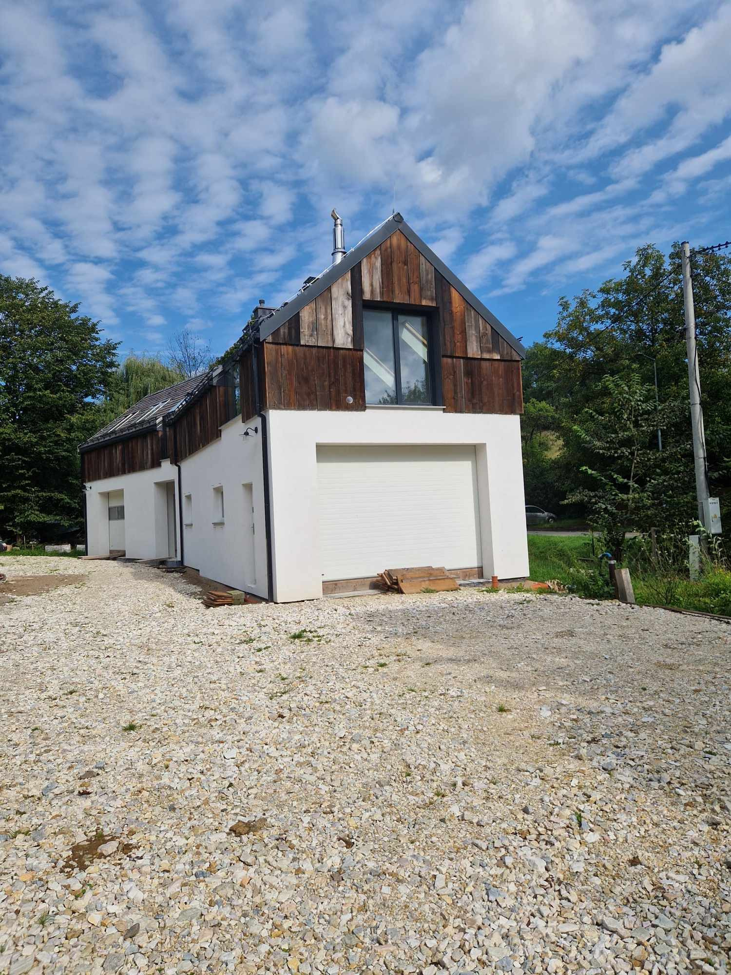 Nowoczesny dom z białą elewacją i drewnianym wykończeniem górnej części, garaż z bramą rolowaną, otoczony żwirowym podjazdem i zielenią.