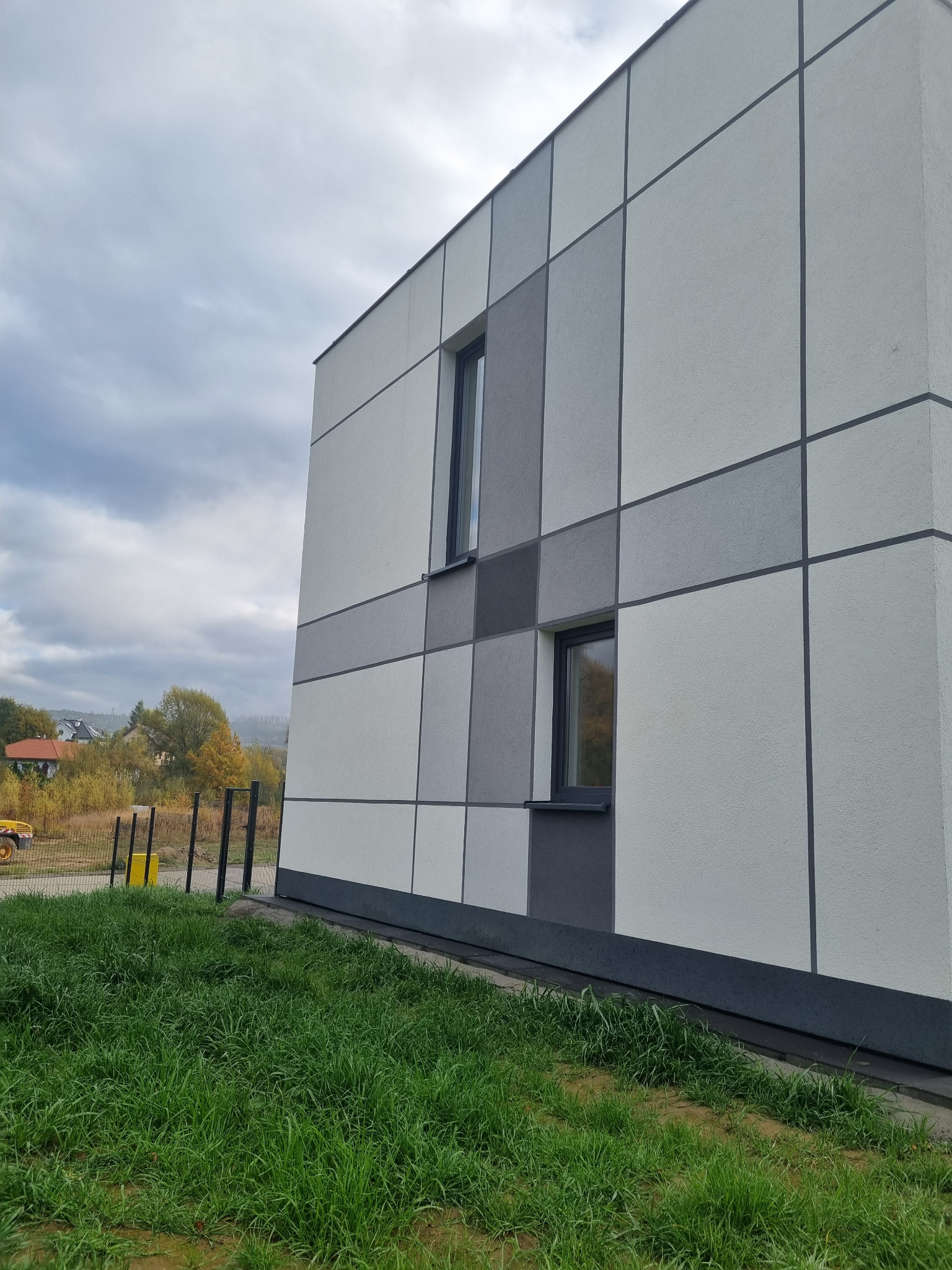 Nowoczesna elewacja budynku z panelami w odcieniach szarości i bieli, widok z dołu, trawa w pierwszym planie, pochmurne niebo w tle.