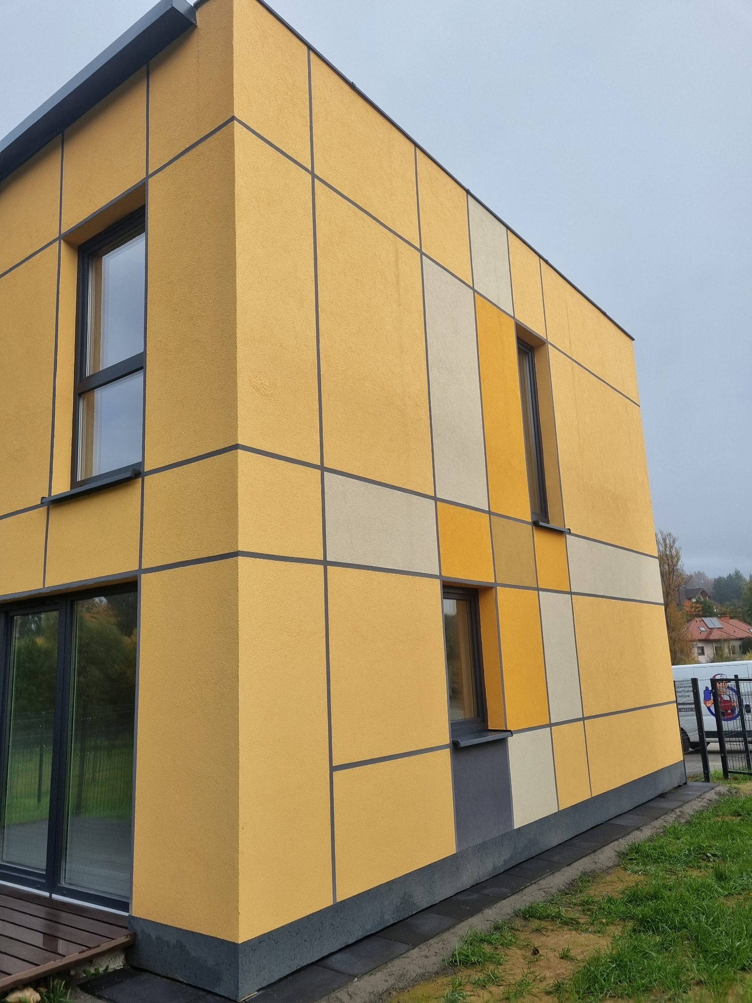 Nowoczesny dom z elewacją w odcieniach żółci i beżu, podzieloną czarnymi listwami na kwadratowe panele, widoczne okna z ciemnymi ramami i fragment tarasu.