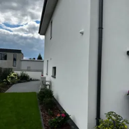 Biała ściana budynku jednorodzinnego z zamontowaną kamerą monitoringu zewnętrznego oraz czarną rurą spustową, widok z boku na tle trawnika i pochmurnego nieba.