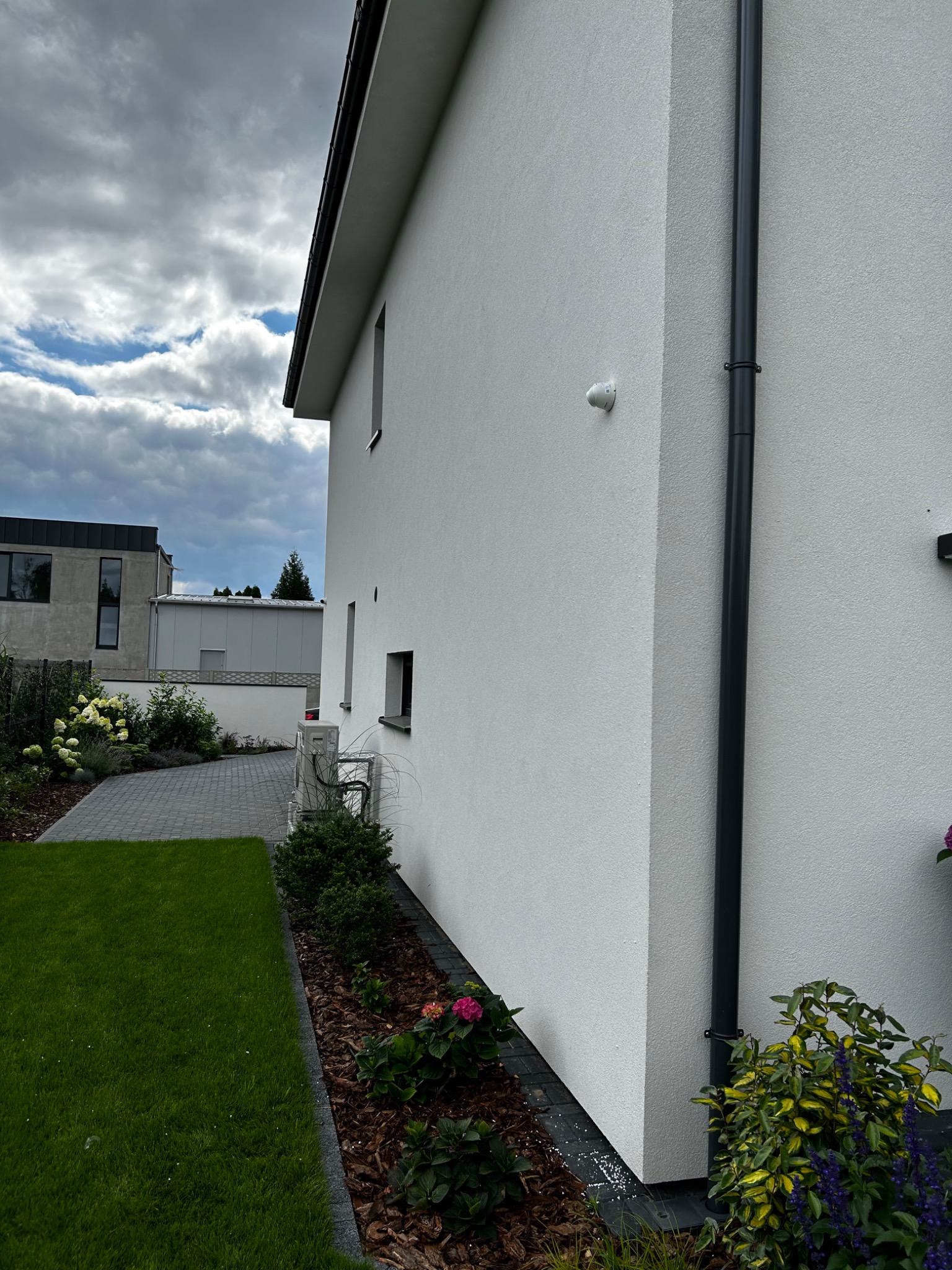 Biała ściana budynku jednorodzinnego z zamontowaną kamerą monitoringu zewnętrznego oraz czarną rurą spustową, widok z boku na tle trawnika i pochmurnego nieba.