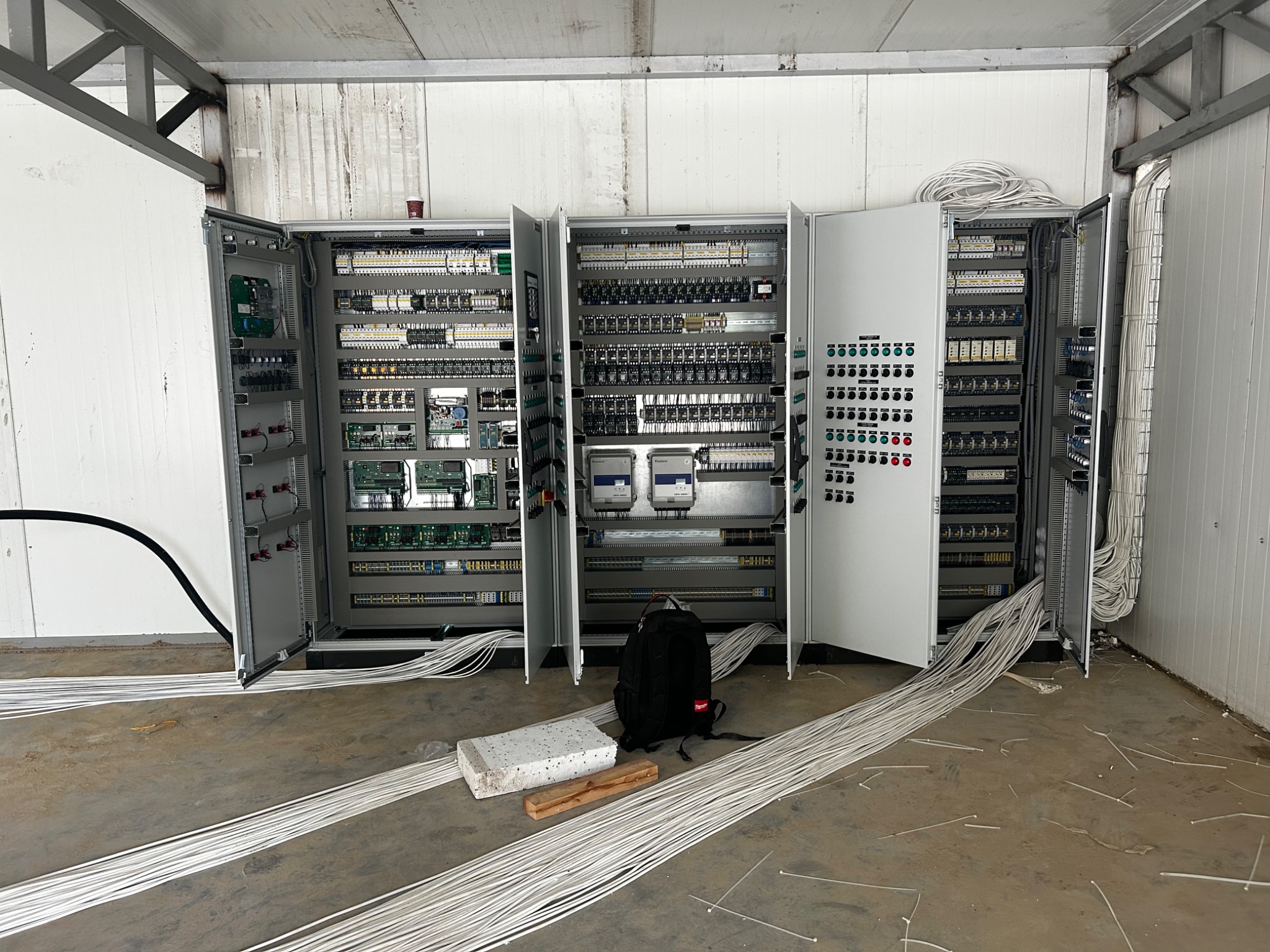 Rozbudowana szafa sterownicza z otwartymi drzwiami, widoczne liczne kable, moduły elektroniczne i przekaźniki, czarny plecak leżący obok, surowy beton na podłodze, białe ściany z paneli.