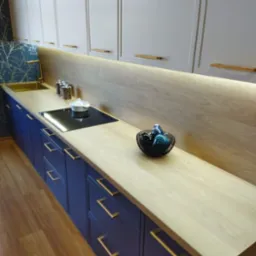 Nowoczesna kuchnia: ciemnoniebieskie szafki z długimi, złotymi uchwytami, jasny drewniany blat, podświetlenie LED pod szafkami wiszącymi, dekoracyjna tapeta z motywem roślinnym.