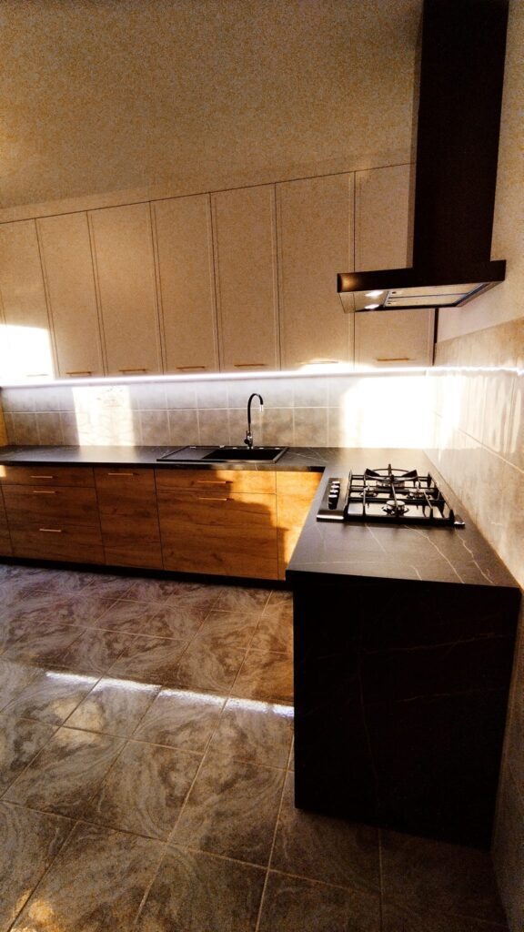 Nowoczesna kuchnia z drewnianymi frontami szafek dolnych, jasnymi szafkami wiszącymi i czarnym blatem z marmurowym wzorem, uzupełniona czarną armaturą i okapem.