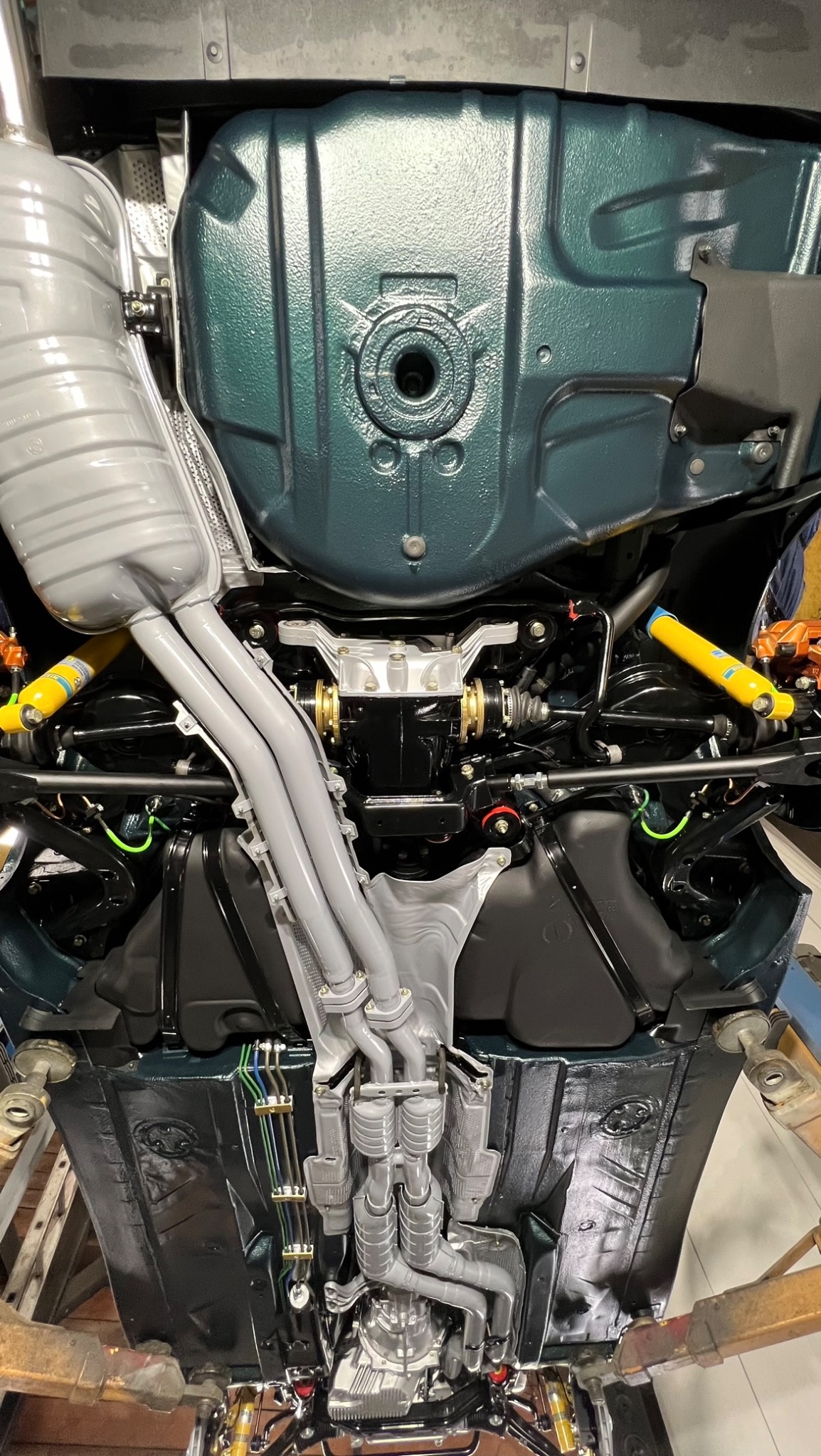 Podwozie samochodu po konserwacji antykorozyjnej, widok od spodu, z elementami układu wydechowego i zawieszenia w odcieniach szarości, czerni i żółci, na podnośniku w warsztacie.