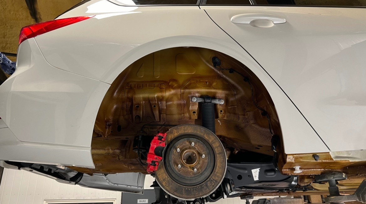 Biały samochód na podnośniku, widoczne zabezpieczone antykorozyjnie nadkole i czerwony zacisk hamulcowy.