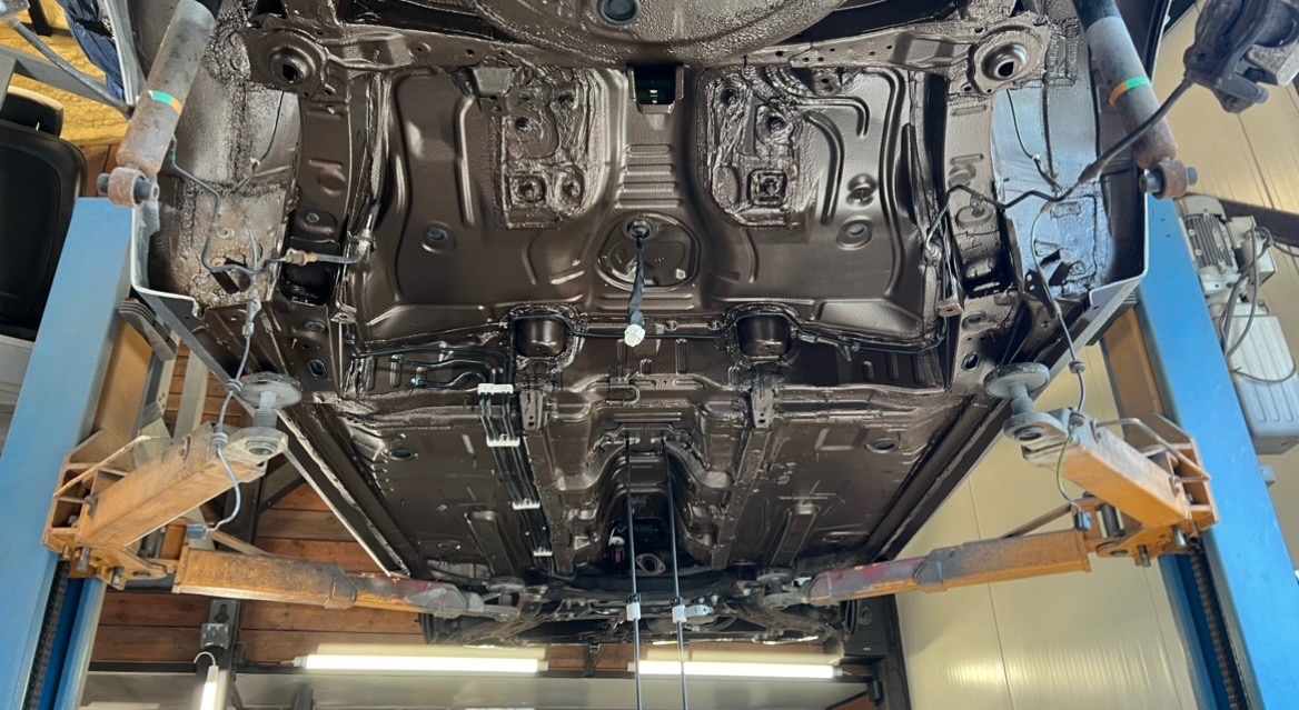 Podwozie samochodu po nałożeniu brązowej powłoki ochronnej, widoczne na podnośniku warsztatowym z perspektywy od dołu.