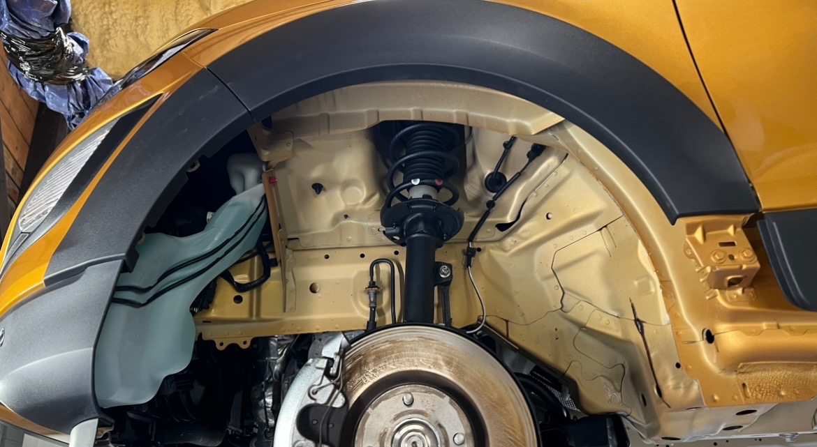 Wnętrze nadkola złotego samochodu z widocznym zawieszeniem, amortyzatorem, sprężyną i zbiornikiem płynu do spryskiwaczy.