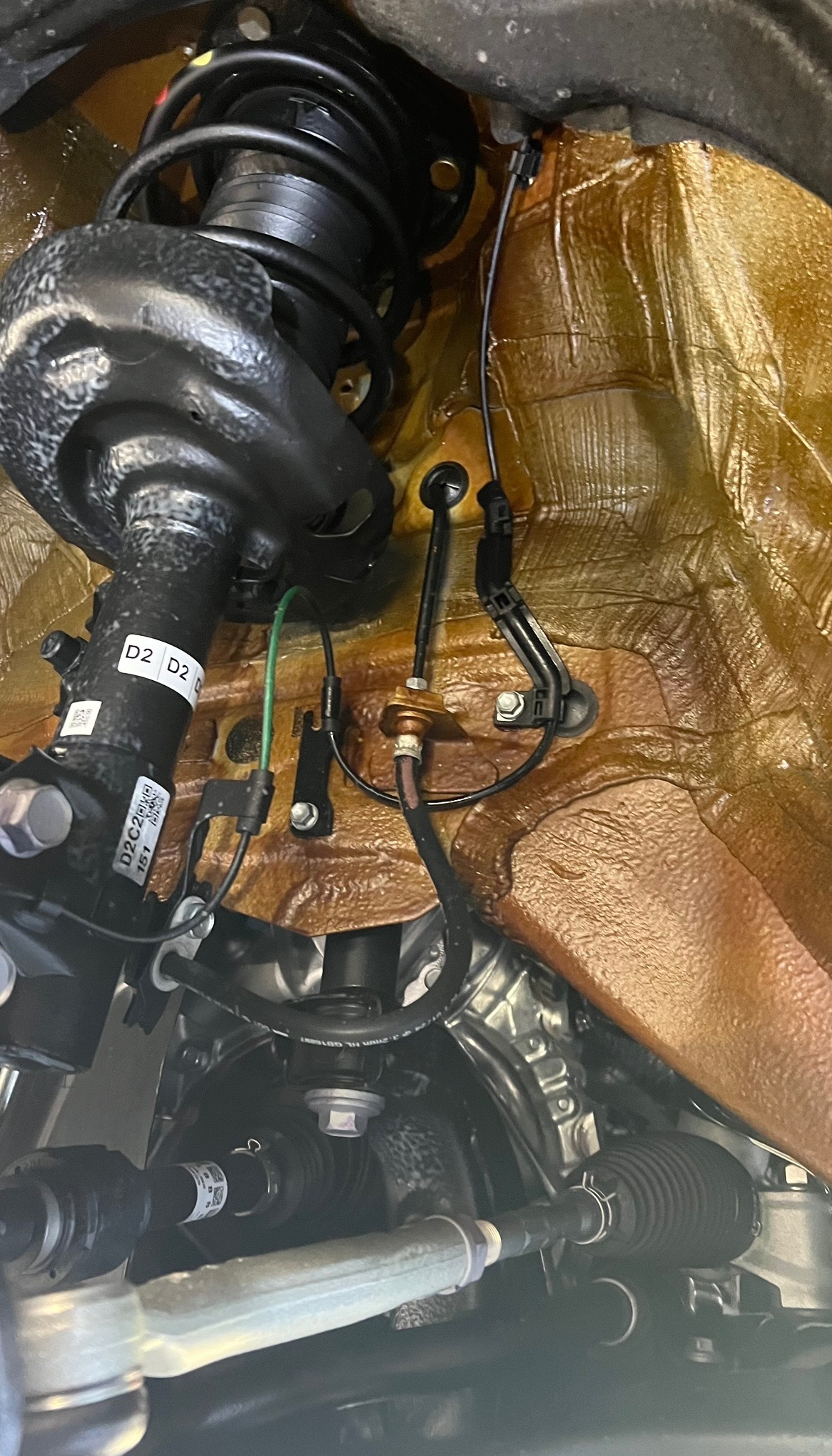 Widok podwozia samochodu z widocznymi elementami zawieszenia, takimi jak amortyzator, sprężyna, przewody i elementy mocujące, z wyraźnie widoczną warstwą ochronną na karoserii w kolorze...