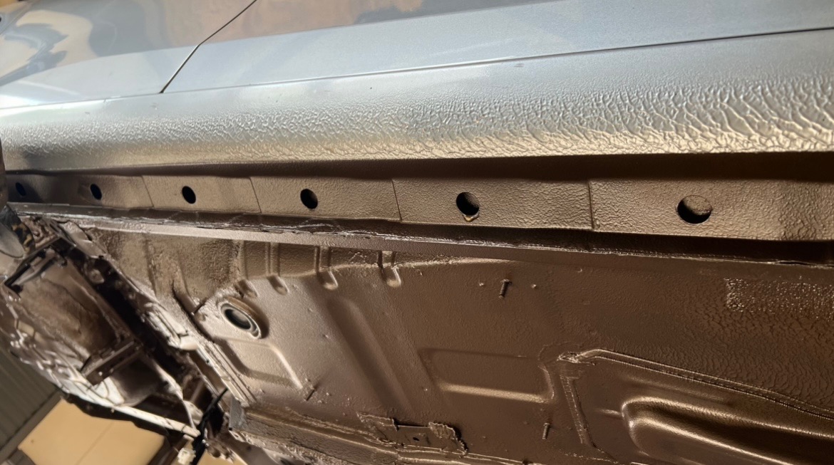 Spód samochodu po zabezpieczeniu antykorozyjnym, widoczne otwory technologiczne i struktura nałożonej powłoki ochronnej w odcieniach brązu.