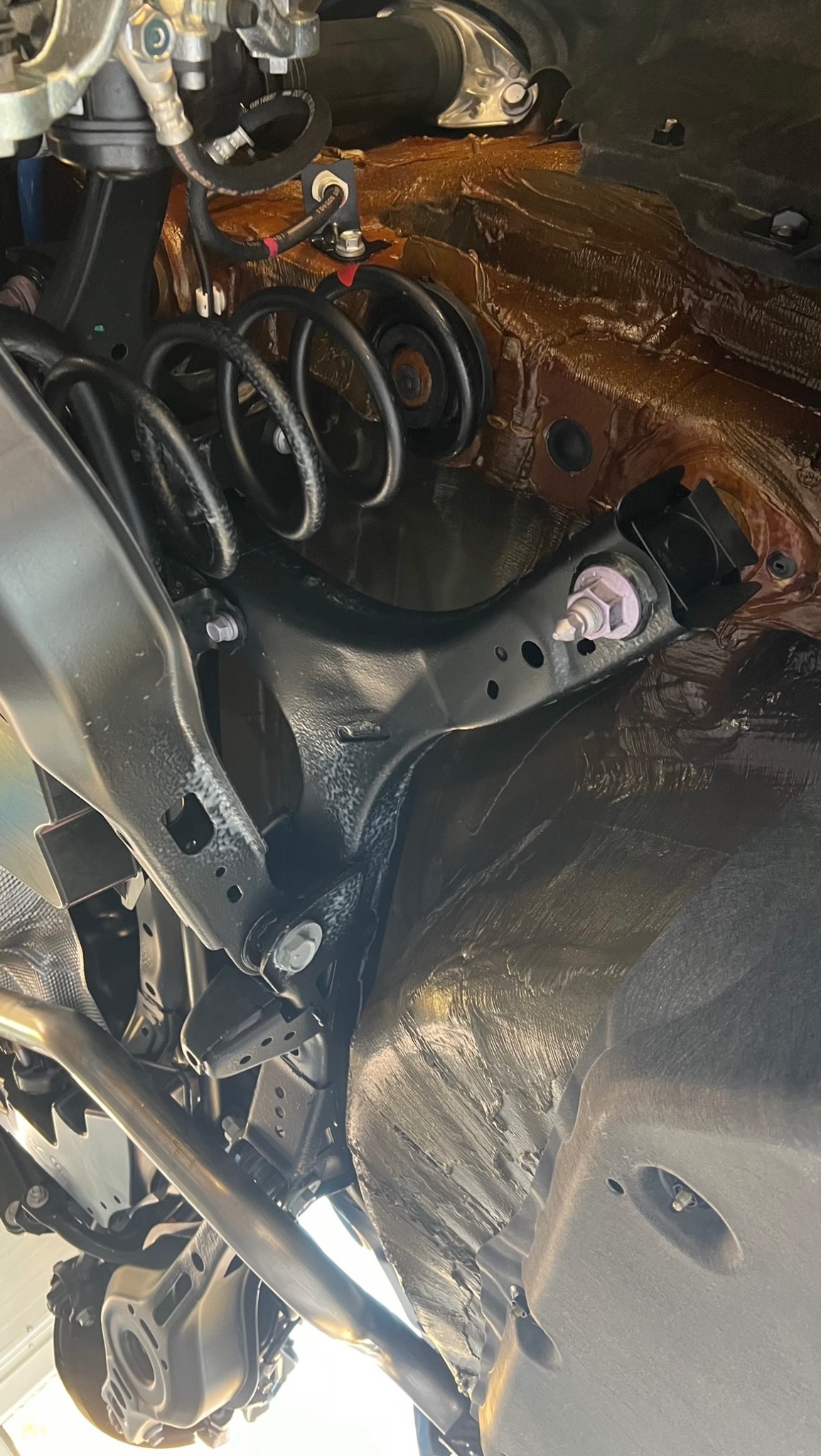 Podwozie samochodu po nałożeniu warstwy ochronnej, widoczne elementy zawieszenia, sprężyny i elementy mocujące. Powierzchnia pokryta gęstym preparatem antykorozyjnym o brązowym zabarwieniu.