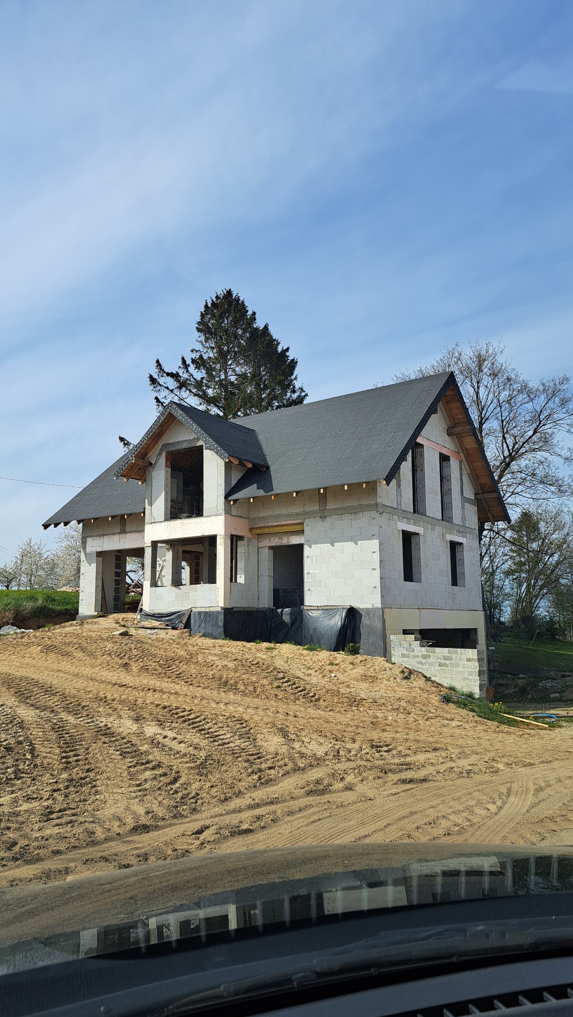 Surowy stan domu z szarej cegły z czarnym dachem na wzgórzu z piaskiem, widok zza szyby samochodu.