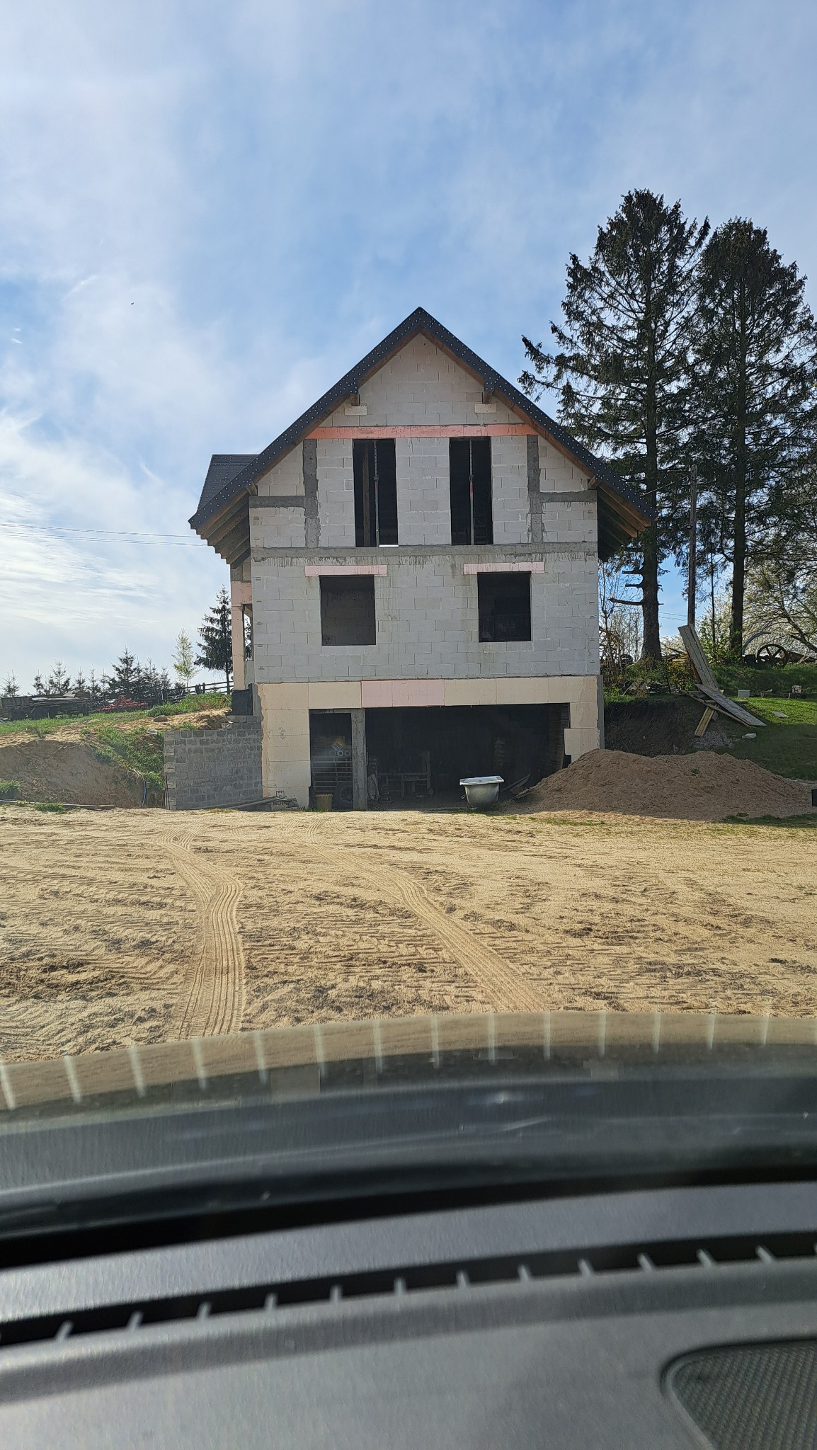 Surowy stan domu w trakcie budowy, ściany z bloczków, widoczne otwory okienne i drzwiowe, dach pokryty dachówką, teren wokół przygotowany pod dalsze prace, widok zza szyby samochodu.