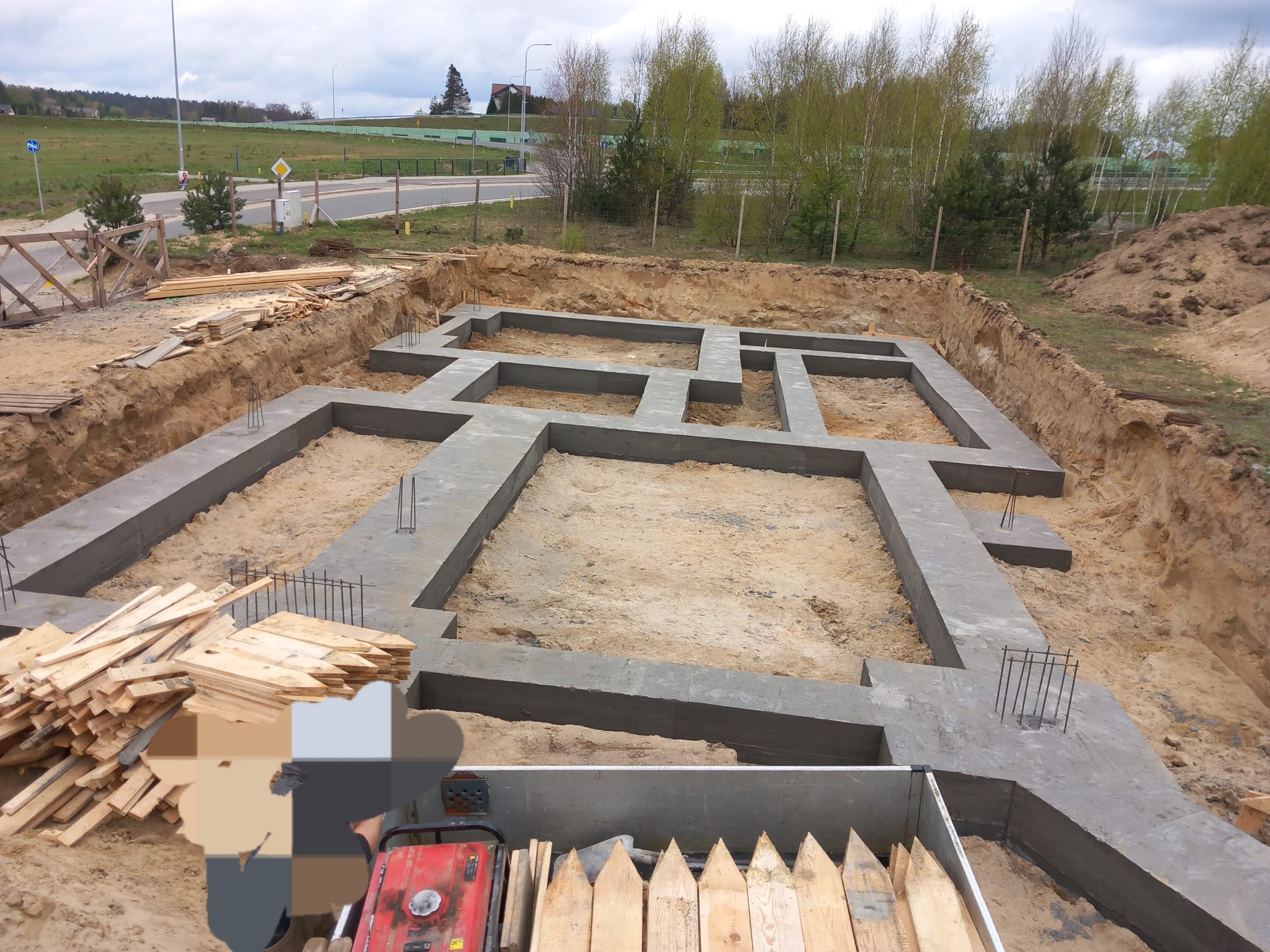 Wykop z betonowymi fundamentami pod budowę domu jednorodzinnego, widoczne wystające pręty zbrojeniowe, deski szalunkowe i agregat prądotwórczy w wykopie, w tle droga i zieleń.