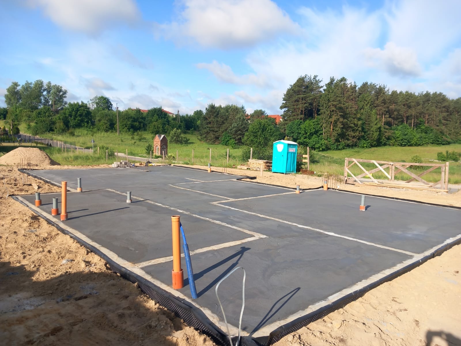 Wylana płyta fundamentowa z wystającymi rurami kanalizacyjnymi, widoczna niebieska toaleta przenośna i drewniane ogrodzenie na tle zieleni.