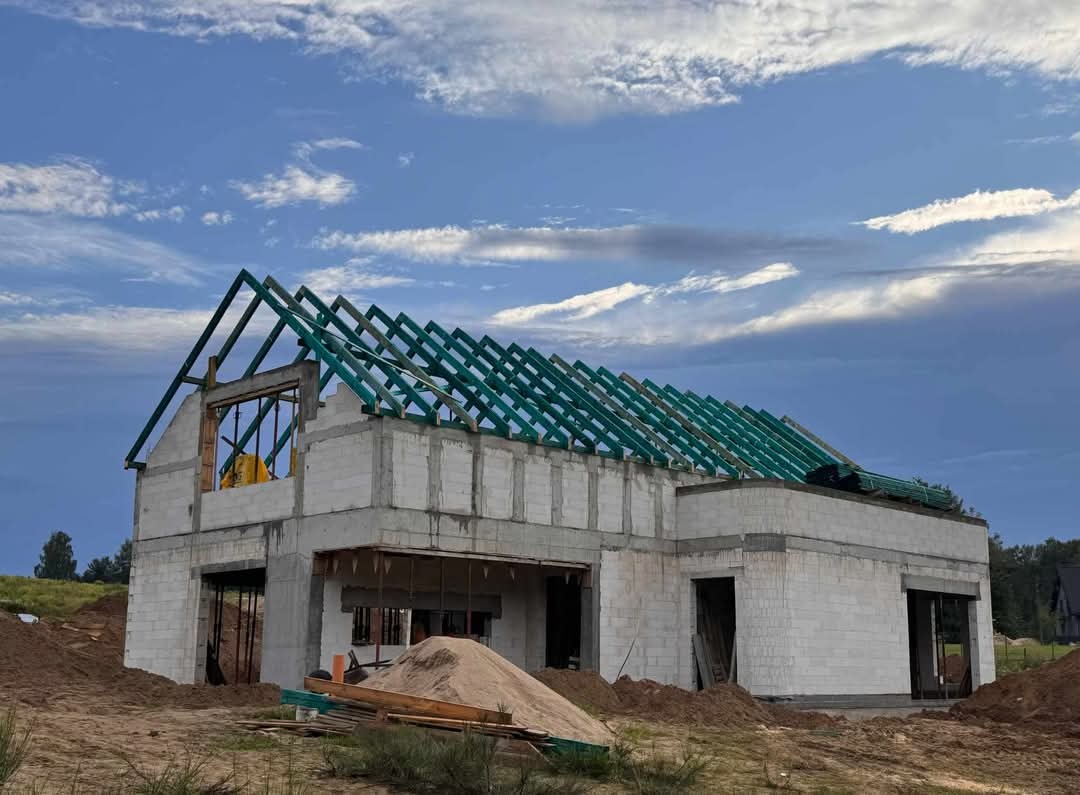 Surowy stan domu jednorodzinnego w trakcie budowy, z widoczną konstrukcją dachu pomalowaną na zielono i ścianami z betonu komórkowego, na tle błękitnego nieba z obłokami.