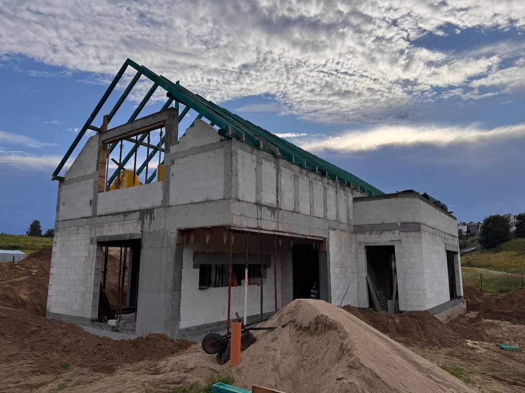 Niedokończony dom z białego betonu komórkowego, widoczne szare spoiny, zielona konstrukcja dachu, sterta piasku i taczka na pierwszym planie, pochmurne niebo w tle.