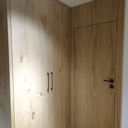 First Floor sp. z o.o - Zabudowa narożna z szafą i drzwiami o identycznym, jasnym wykończeniu drewnopodobnym, z czarnymi, minimalistycznymi uchwytami i klamką.